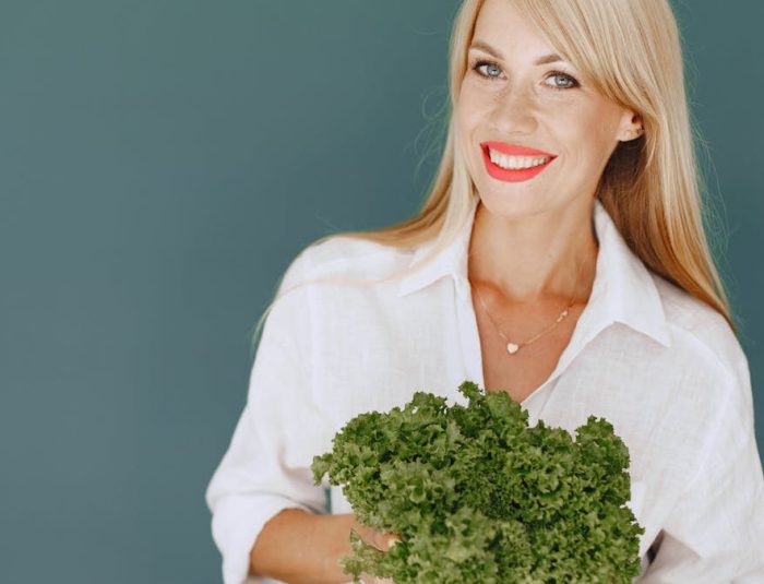 Cheerful woman with blonde hair holding fresh lettuce, promoting healthy living in a studio setting.