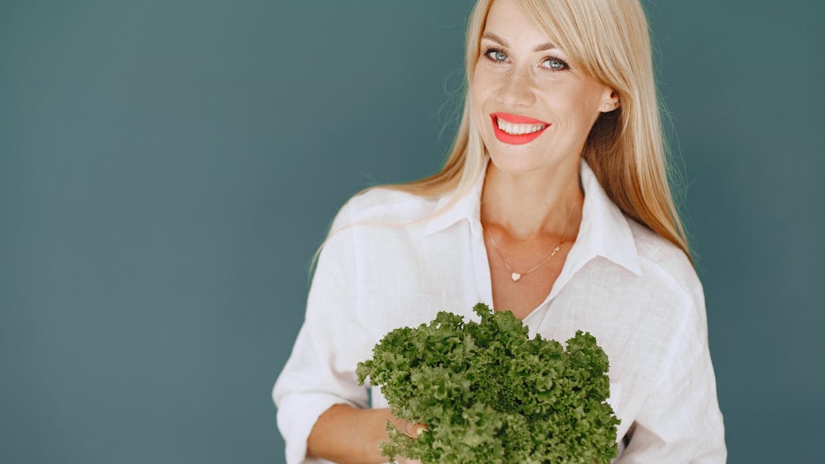 Cheerful woman with blonde hair holding fresh lettuce, promoting healthy living in a studio setting.
