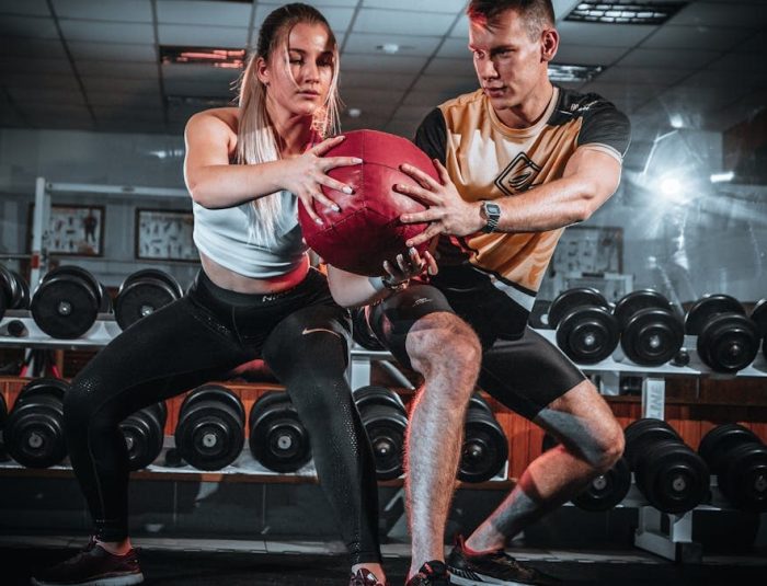 A man and woman perform a tandem workout with a medicine ball in a gym setting.